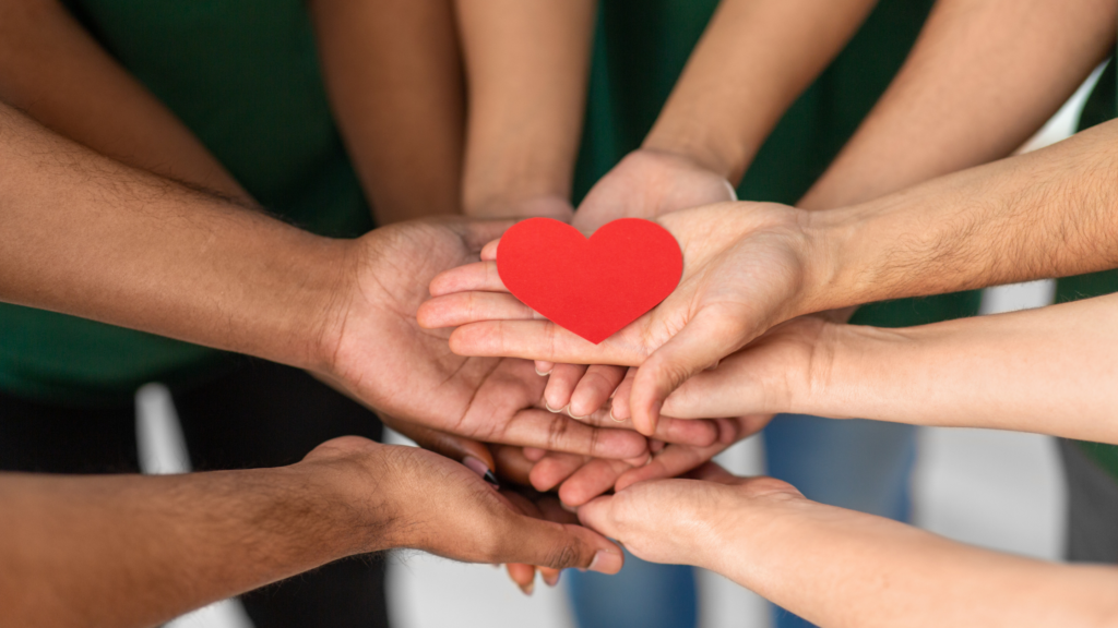 Hands and arms are reaching into the center of a circle of people. The hands are all supporting red paper heart.