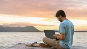 Digital nomad sits on a dock at sunset working on a laptop