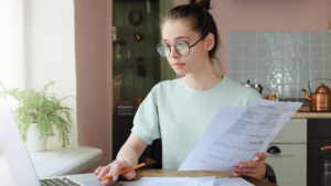 A woman wearing glasses works at her laptop near a bright window